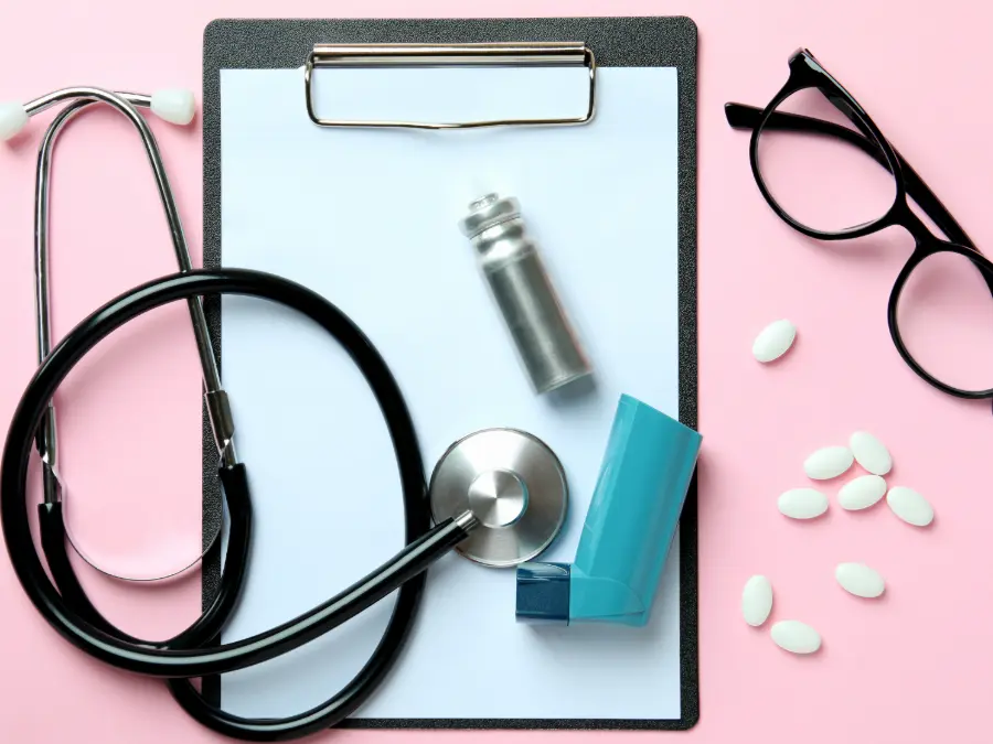 Doctors stethscope, an inhaler, glasses, pills, a clipboard all on a pink table.